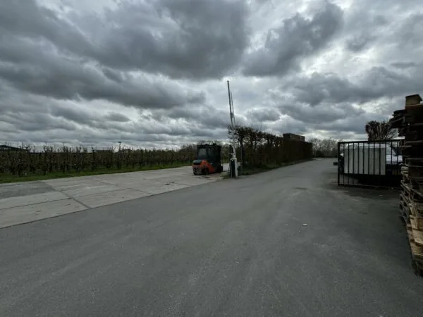 Een geasfalteerde weg met een heftruck en slagboom aan de Graaf van Lynden van Sandenburgweg, omringd door boomgaarden en stapels pallets onder een bewolkte lucht.